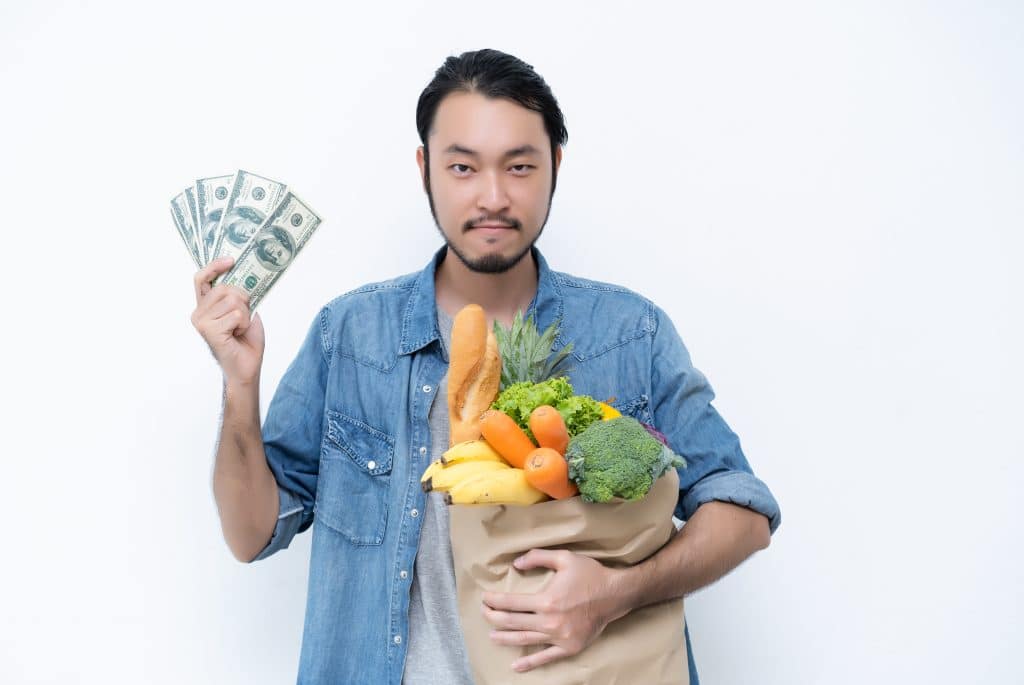 Asian male shopper, hands holding shopping bags full of groceries and show dollar bills, isolated standing on white background.