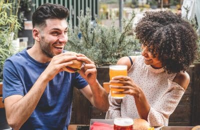 young couple having fun at restaurant, two diverse friends eating burgers at fast food and drinking beer